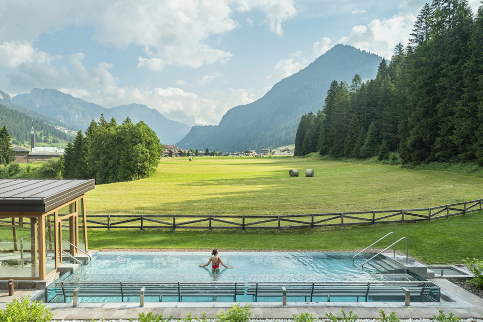 A person relaxing in an outdoor pool overlooking a scenic meadow with mountains and trees in the background on a sunny day.