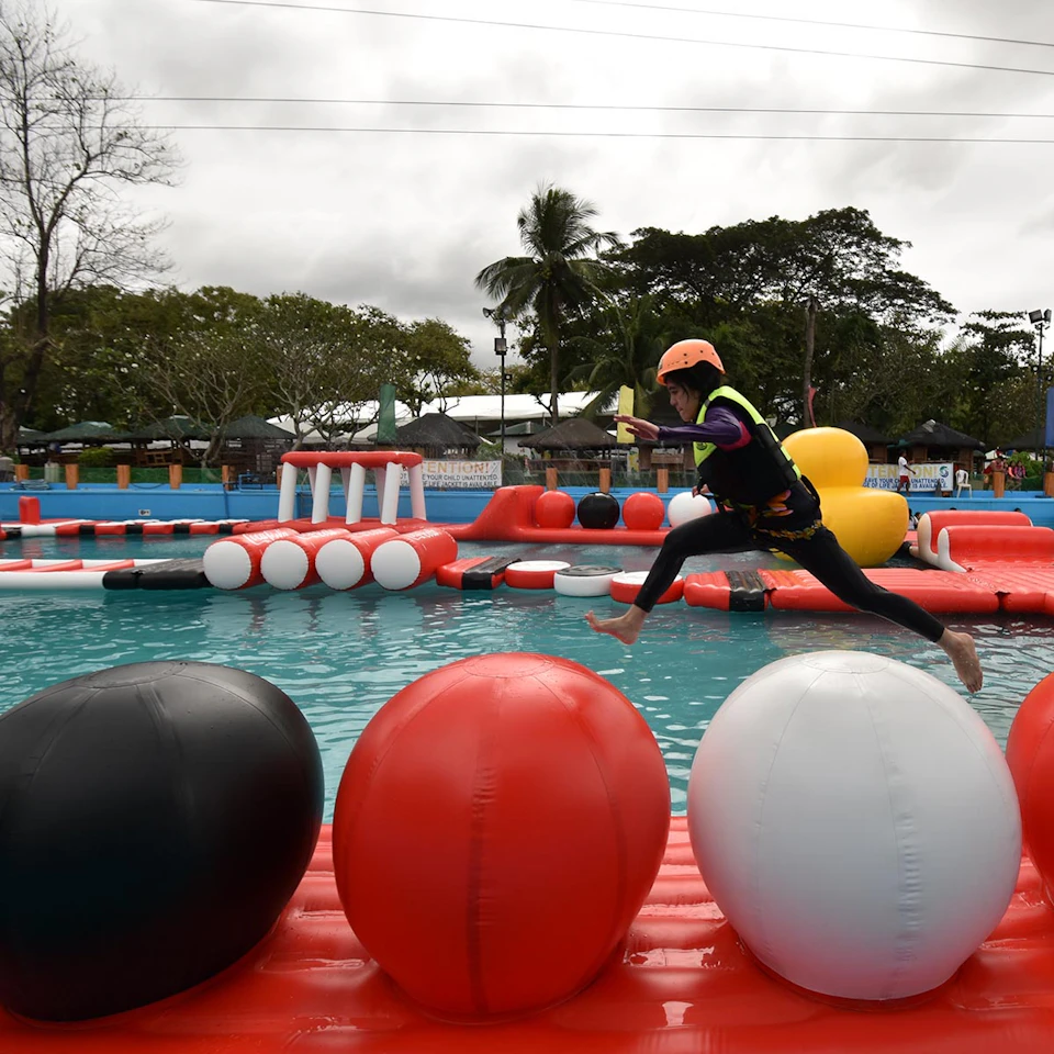 A person wearing a helmet and safety gear leaps over large inflatable balls on a water obstacle course in an outdoor setting.