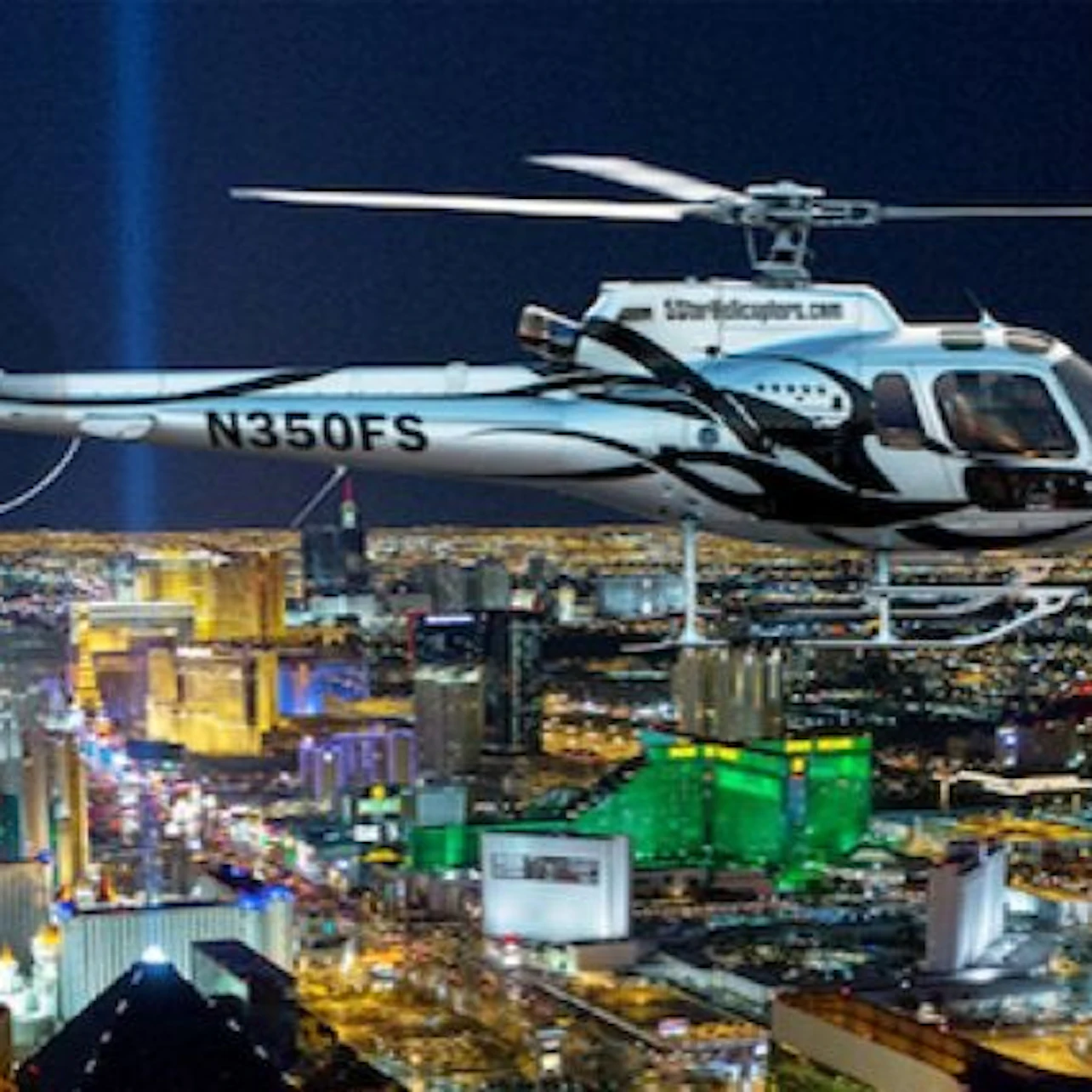 A helicopter flying over a brightly lit cityscape at night, with prominent city lights and buildings below.