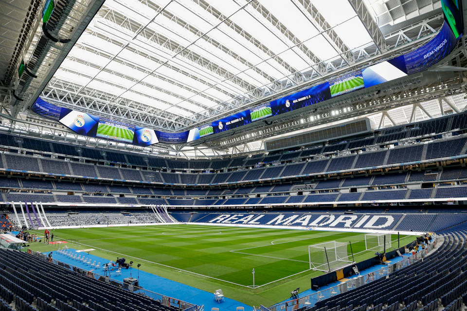 An empty football stadium with a green field, stands displaying 'Real Madrid,' and a partially closed roof.
