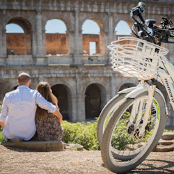 Ett par sitter omfamnande nära Colosseum, med en vit cykel med en främre korg i förgrunden.