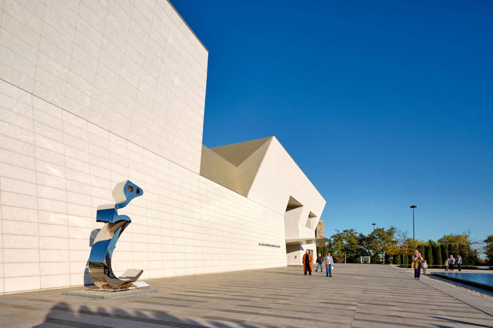 Modern white building with angular design, a reflective sculpture in the foreground, and people walking nearby.