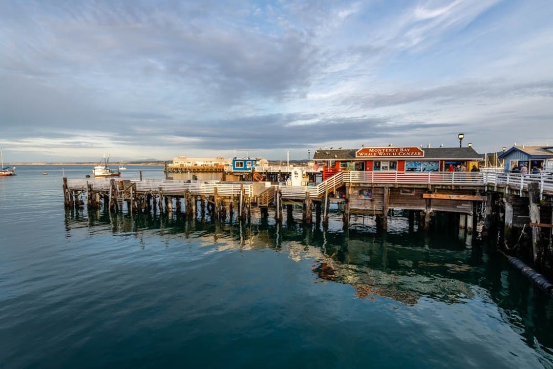 Wooden pier with various buildings overlooking calm water. Sign reads "Monterey Bay Whale Watch Center." Overcast sky.