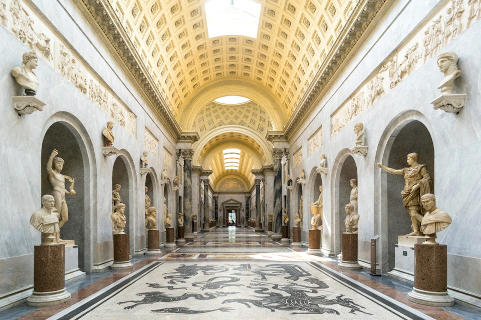 A grand hall with a vaulted ceiling, classical statues lining the walls, and intricate artwork on the floor.