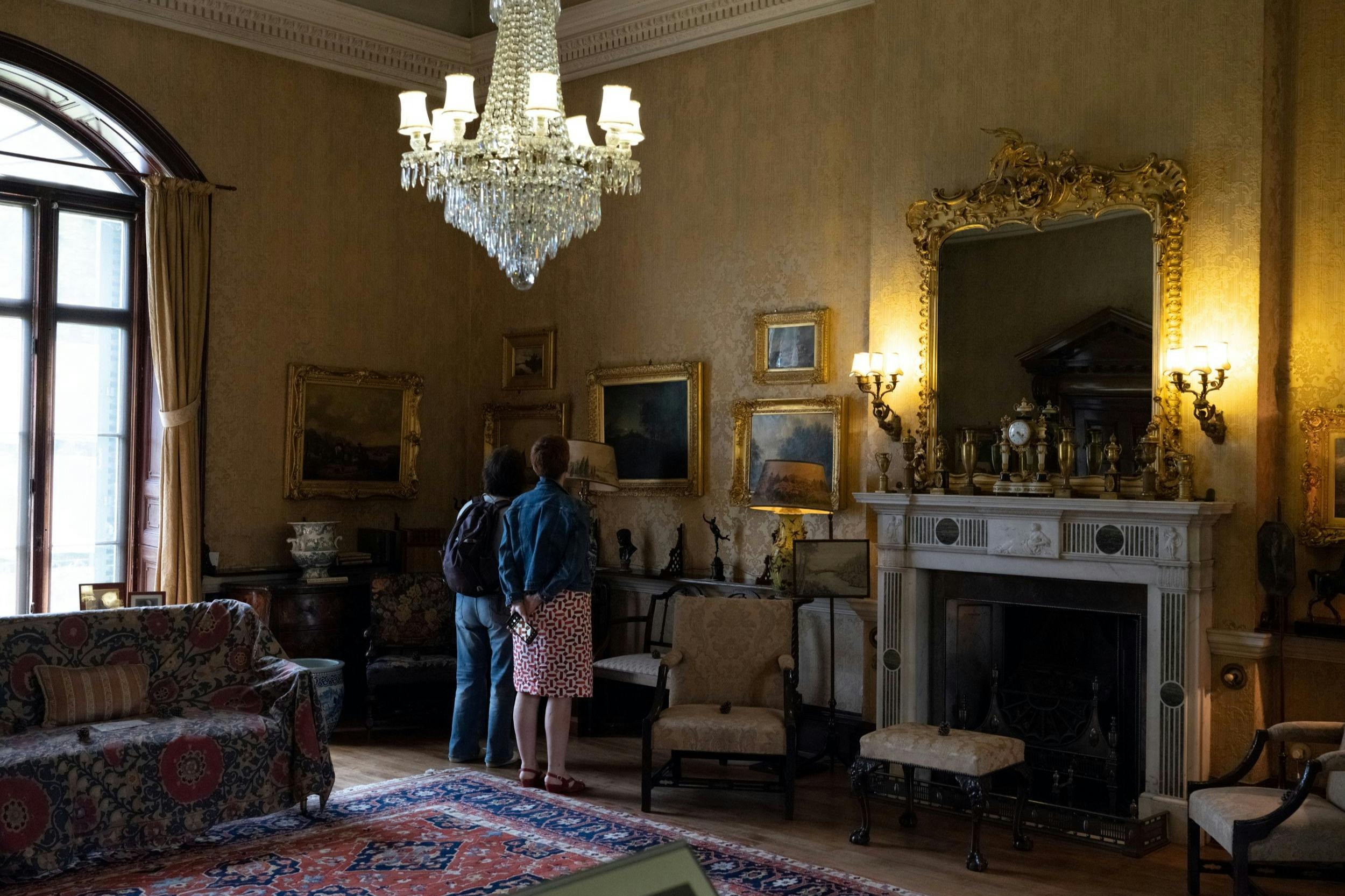 Zwei Personen betrachten Gemälde in einem prunkvollen Raum mit Kronleuchter, Kamin, antiken Möbeln und einem großen Spiegel.