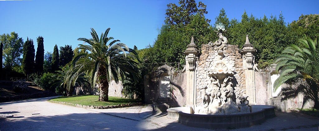 Une fontaine en pierre avec des sculptures dans un jardin, entourée de palmiers et de verdure sous un ciel bleu clair.