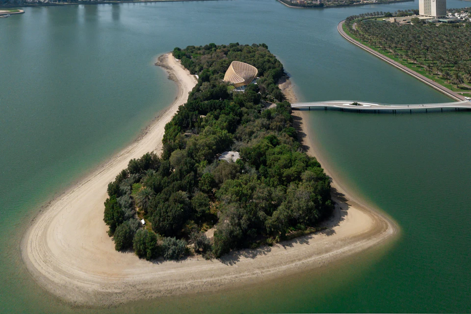 Aerial view of a lush, forested island with a dome-shaped building, sandy shores, and a bridge connecting to the mainland.