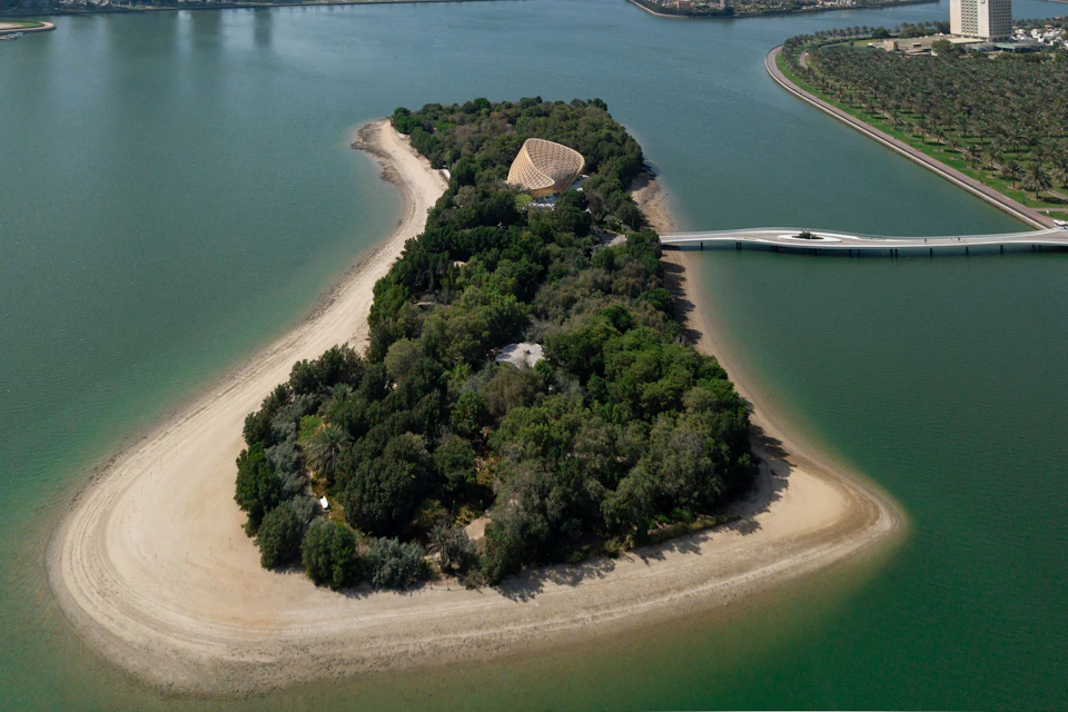 Aerial view of a lush, forested island with a dome-shaped building, sandy shores, and a bridge connecting to the mainland.