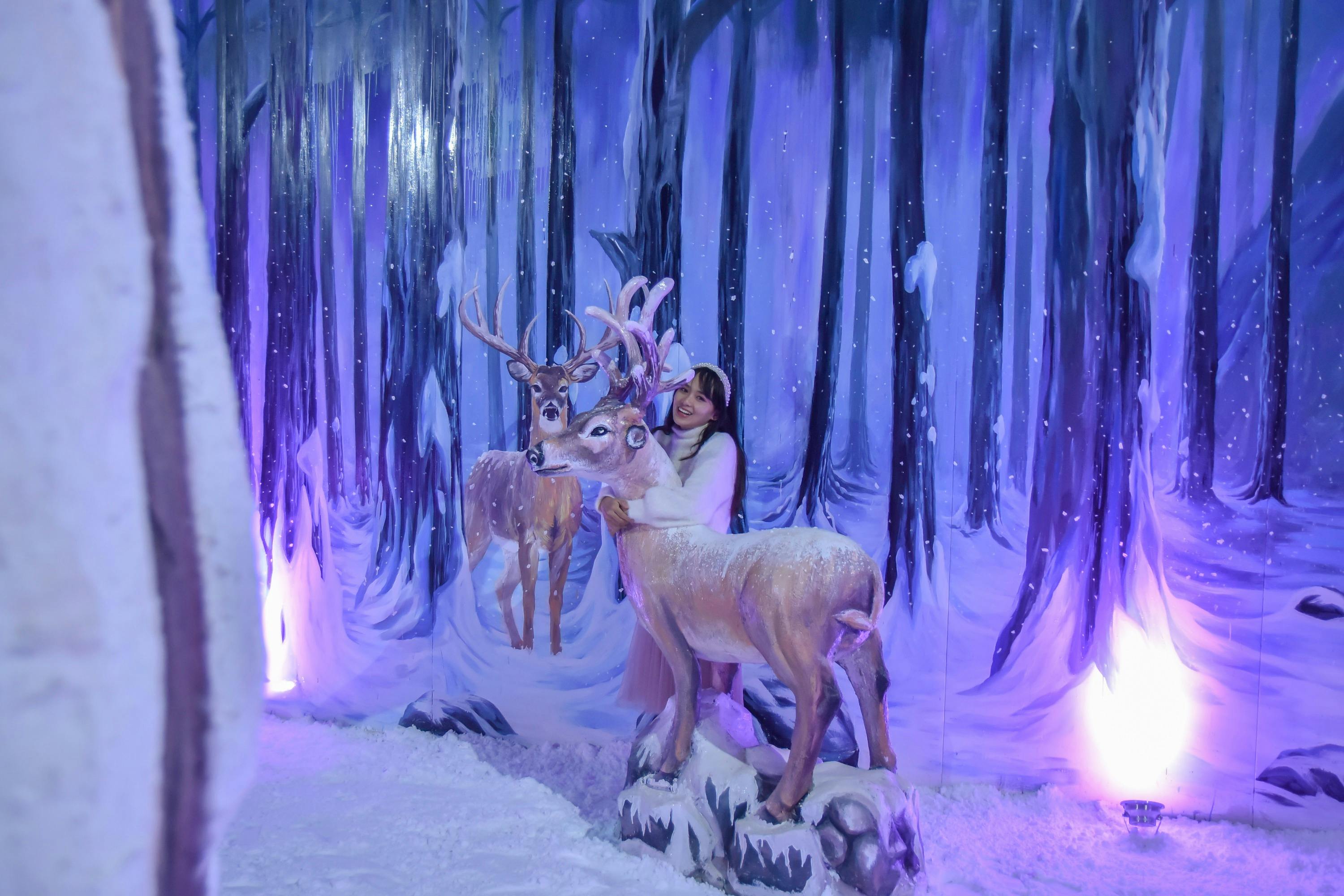 A person posing with a deer statue in a snowy, forest-themed setting, with painted trees and winter decorations.