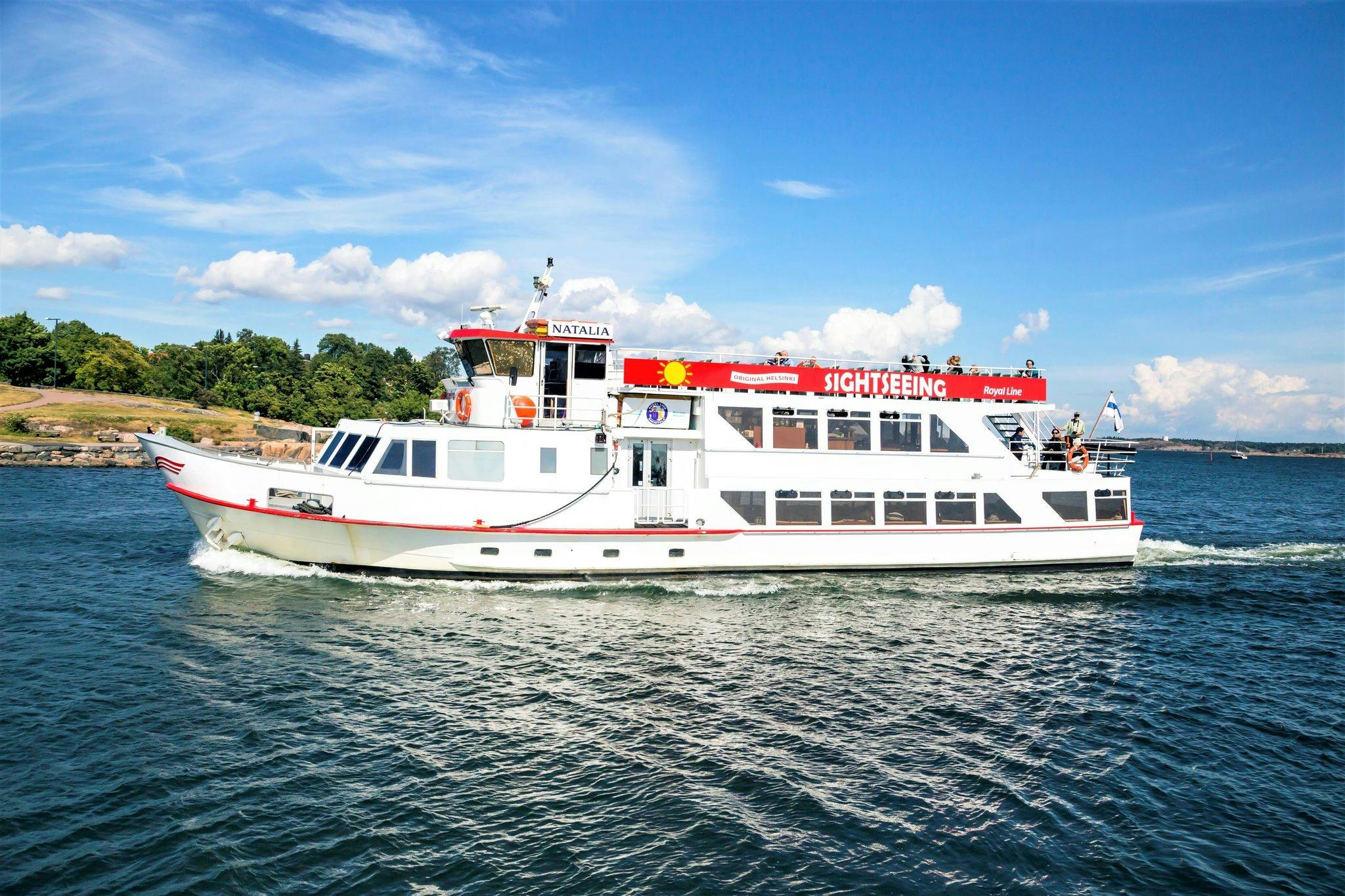 A white sightseeing boat named Natalia cruising on water near a shoreline with trees under a partly cloudy blue sky.