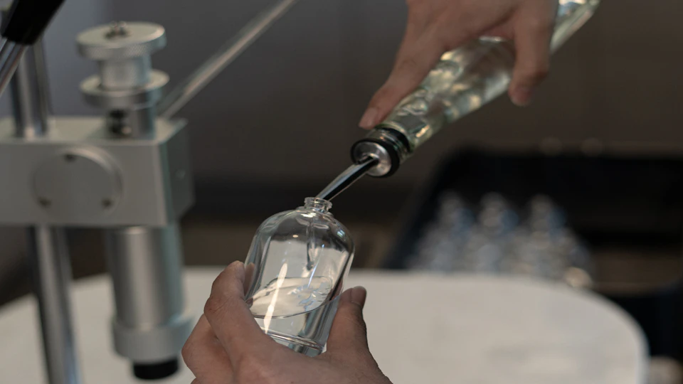 A close-up of hands filling a small glass bottle with liquid using a spout, with blurred background equipment visible.