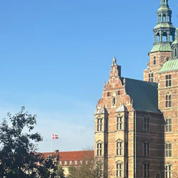 Historic castle with ornate towers and a red brick facade under a clear blue sky; a Danish flag flies in the background.