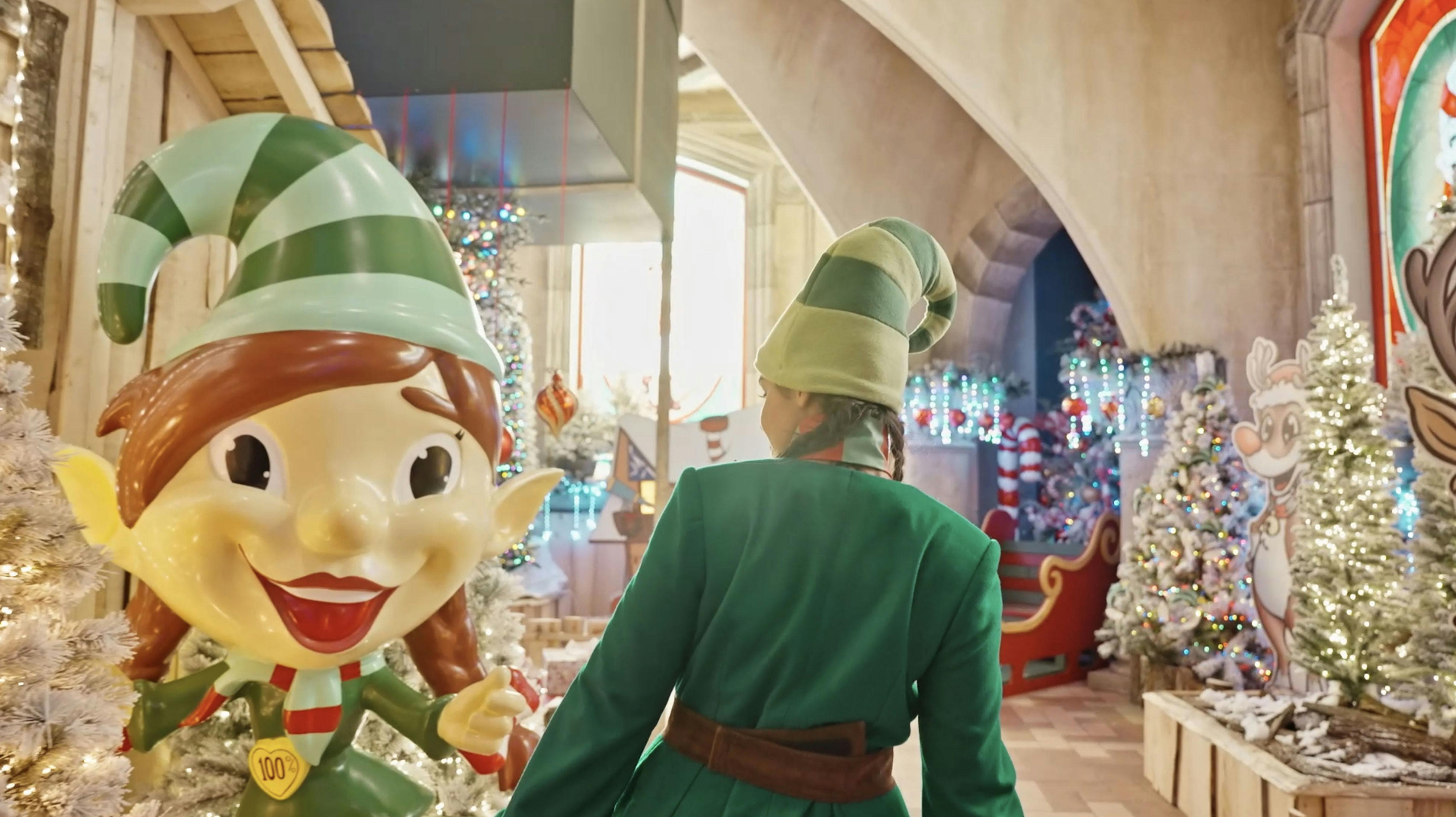 초록색 엘프 의상을 입은 사람이 커다란 엘프 동상과 연말연시 불빛으로 장식된 축제 공간에 서 있습니다.