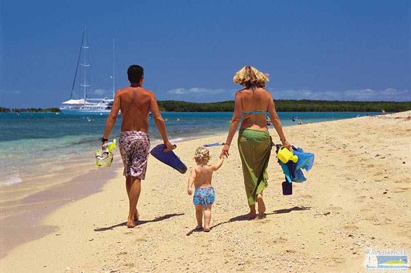 A couple and a child walk along a sandy beach towards the water, carrying snorkeling gear. Sailboats are visible in the background.