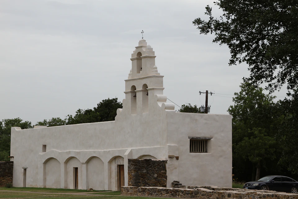 A historic white stucco church with arched doorways, a stone base, and a bell tower, set against a cloudy sky with surrounding greenery.