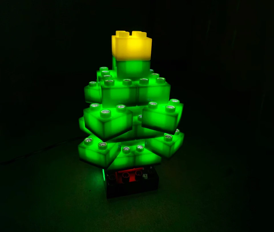 A Christmas tree built with large glowing LEGO-like blocks, primarily green with a yellow top, on a dark background.