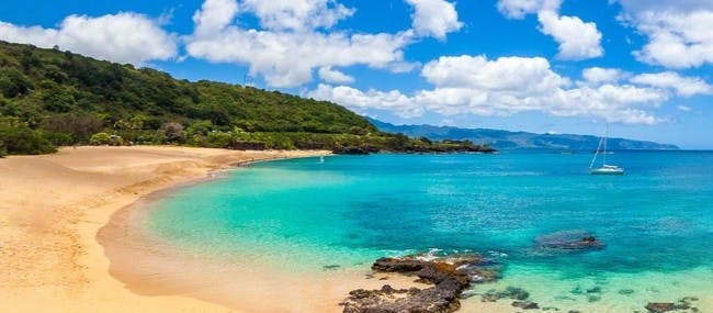 Uma praia tropical com areia dourada, água azul, costa rochosa e colinas verdejantes sob um céu parcialmente nublado.