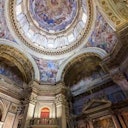 Ornate cathedral interior with detailed frescoes on domed ceilings and archways, showcasing religious scenes and figures.