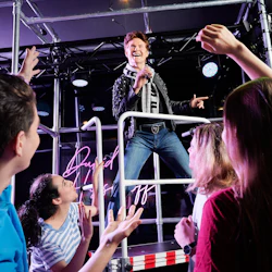 En artist på scenen håller en mikrofon och sjunger som en grupp entusiastiska fans med upplyfta armar under.