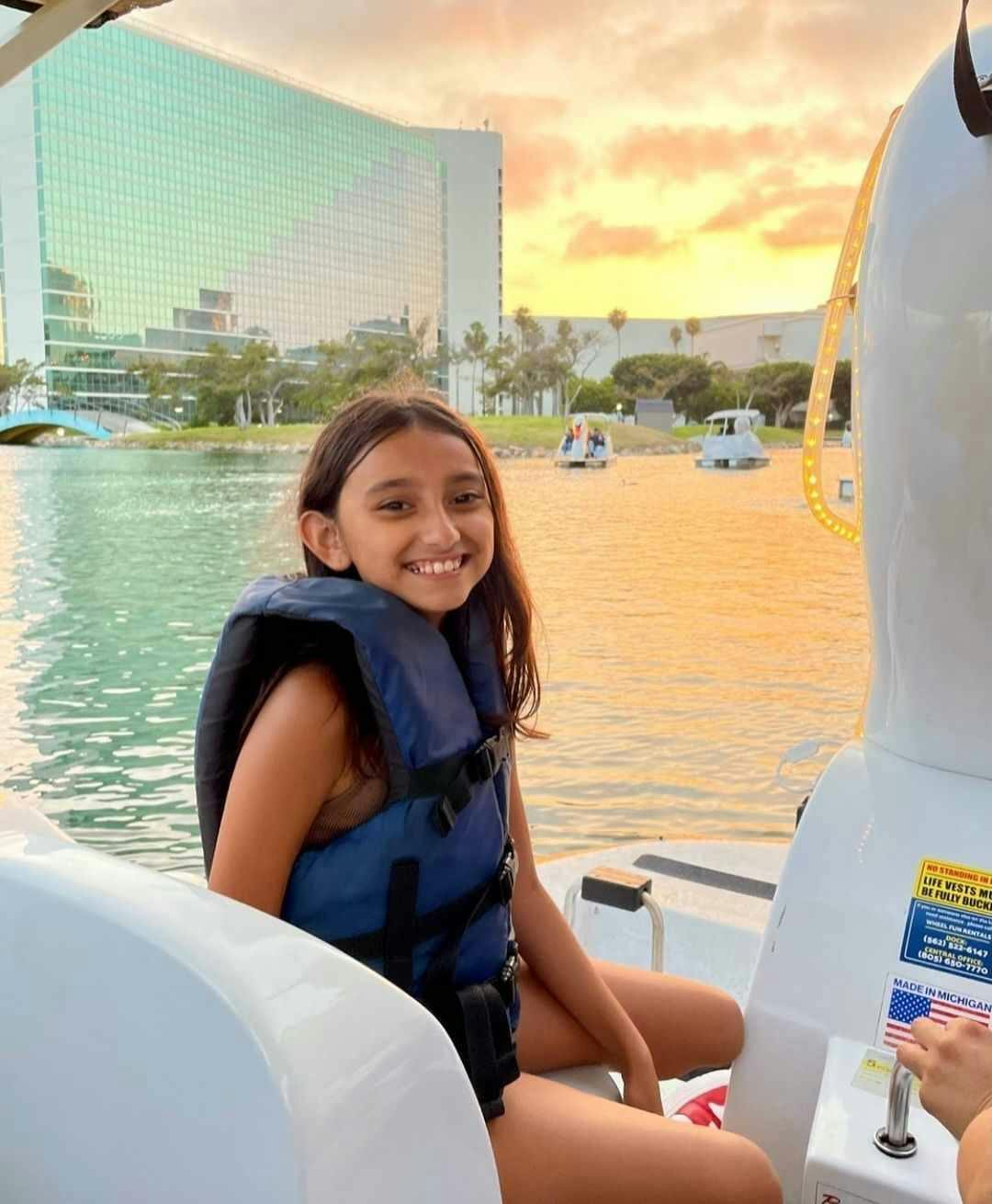 Usmívající se dívka v záchranné vestě na lodi s moderní budovou a západem slunce v pozadí.