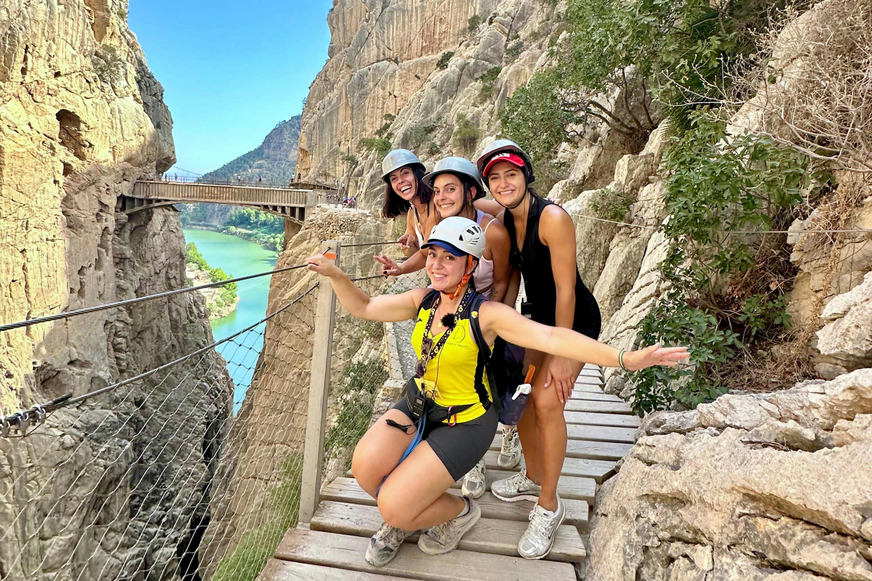 Four people in helmets pose on a narrow wooden pathway along a rocky cliff with a bridge and river in the background.