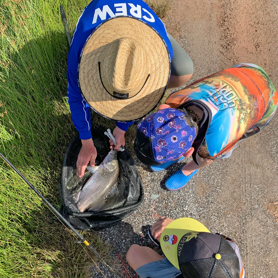 Three people examine a large fish in a net on the ground near a grassy area. One wears a hat and "CREW" shirt, the others wear caps.