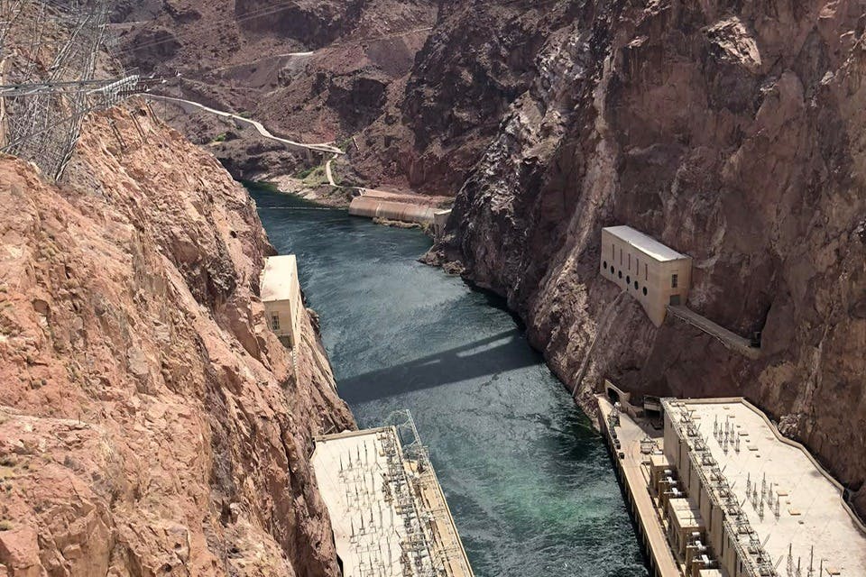 A river flows between steep rocky cliffs, with buildings on both sides of the water, likely part of a dam or hydroelectric plant.