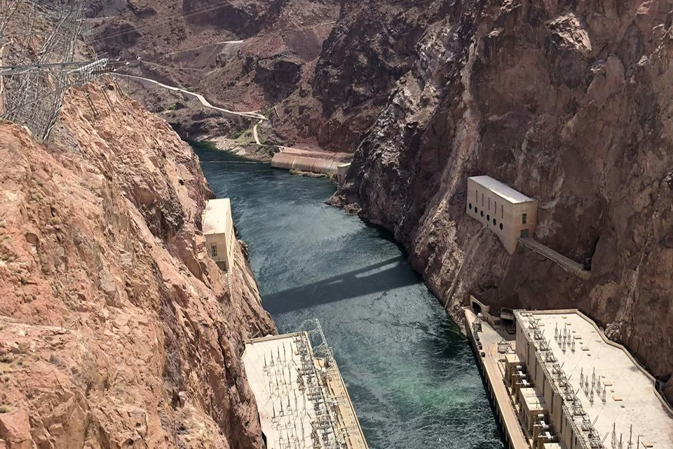 A river flows between steep rocky cliffs, with buildings on both sides of the water, likely part of a dam or hydroelectric plant.