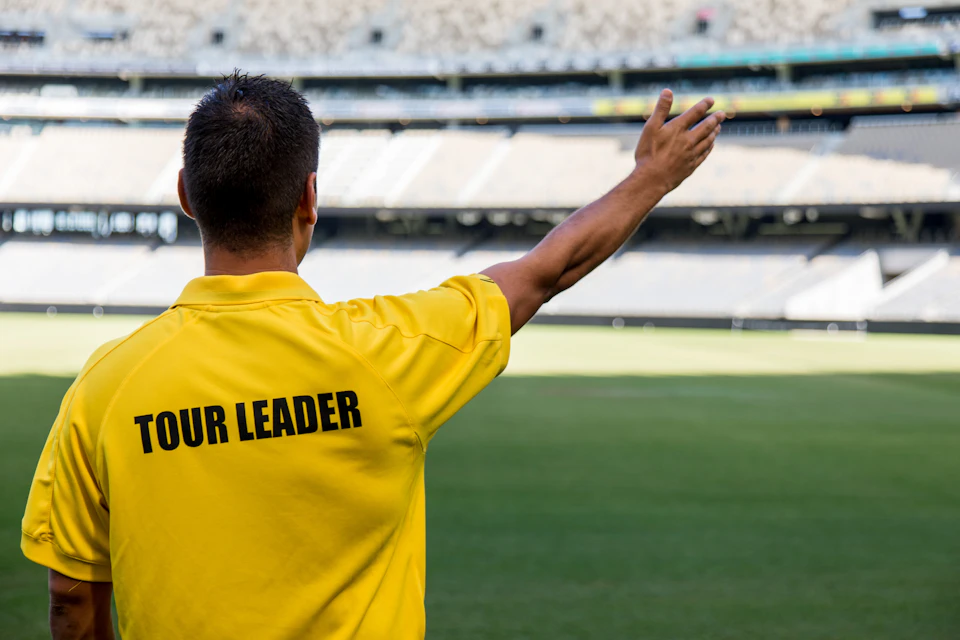 A person in a yellow shirt labeled "TOUR LEADER" gestures with one arm extended, standing in an empty sports stadium.