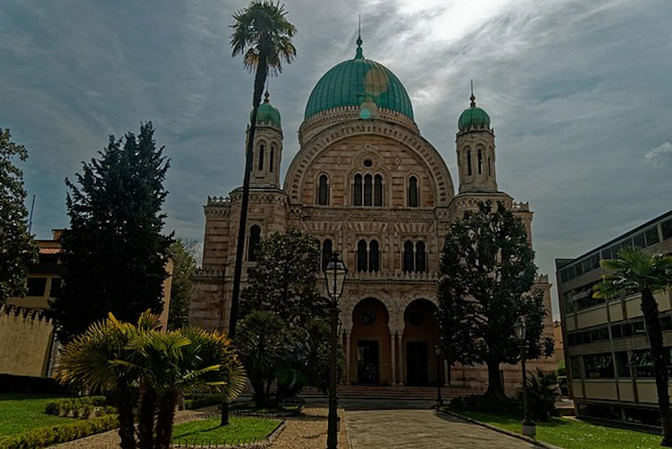 A historical building with a large green dome, flanked by two smaller domes, surrounded by trees and a path leading to the entrance.