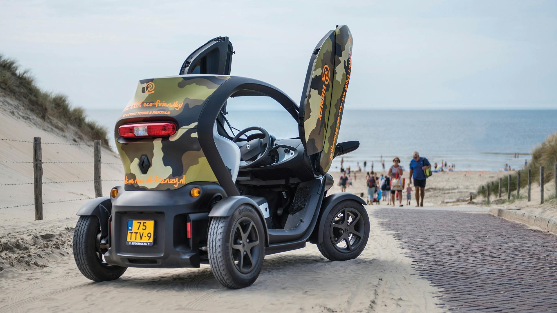 A small camouflage-painted electric vehicle with open doors parked on a sandy path near a busy beach.