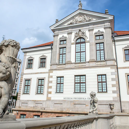 Utsmyckad vit byggnad med välvda fönster och statyer, med texten "Muzeum Fryderyka Chopina", under en klar himmel.