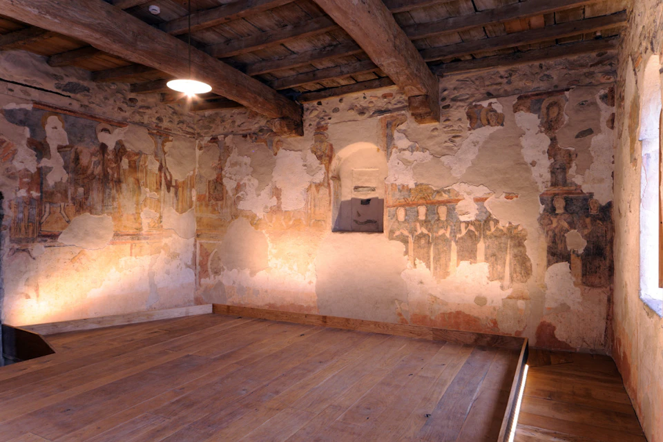 A rustic room with wooden beams, worn fresco paintings on plaster walls, wooden flooring, and a single hanging light fixture.