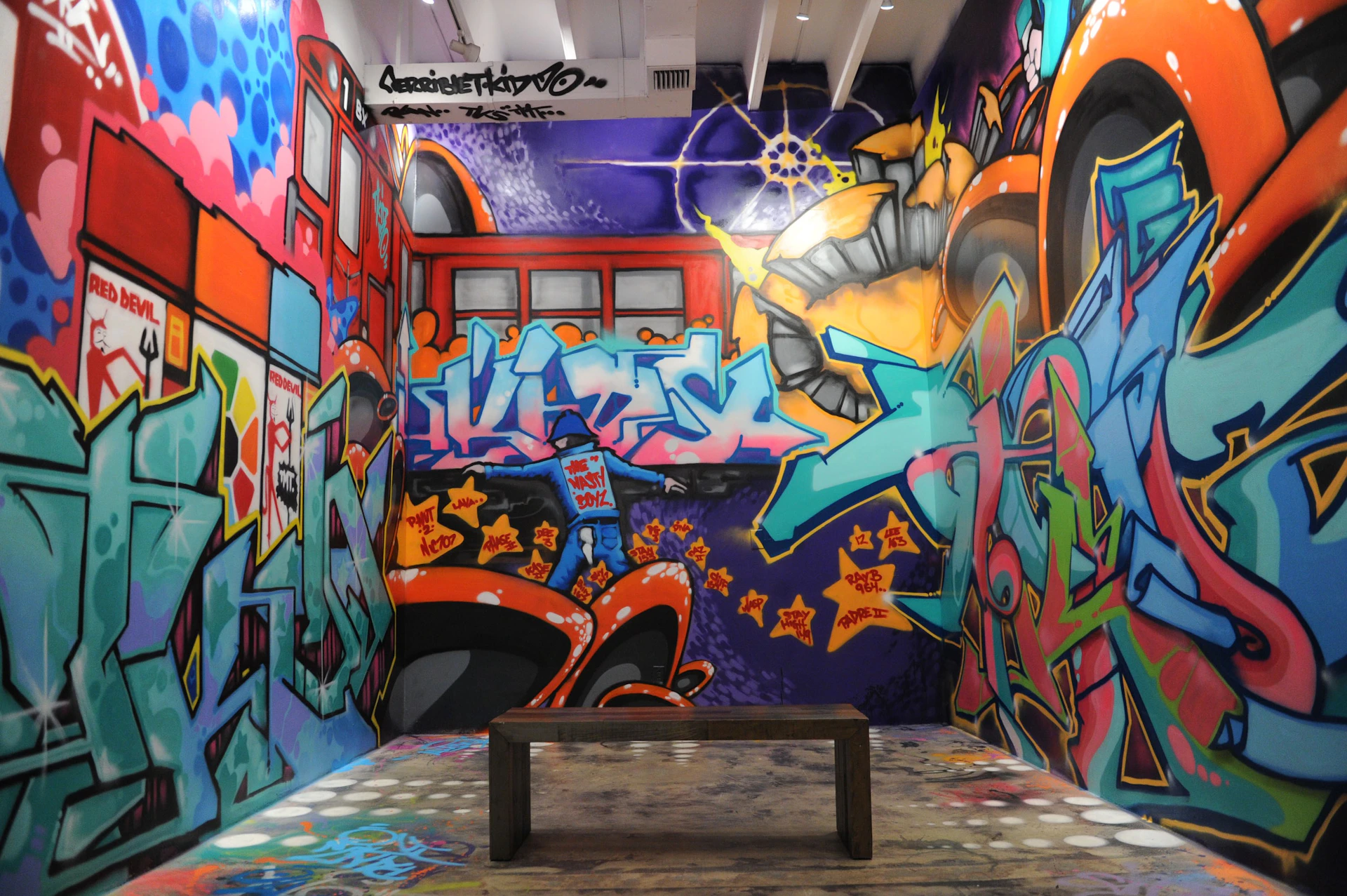 Room covered in colorful graffiti art featuring abstract designs and shapes, with a wooden bench in the center on a worn floor.