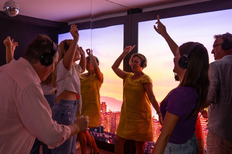 People wearing headphones are dancing enthusiastically in a room with large windows showing a cityscape at sunset.