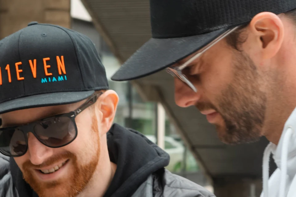 Two men in sunglasses and caps, one in a black jacket and the other in a white hoodie, look at a paper in an urban setting.