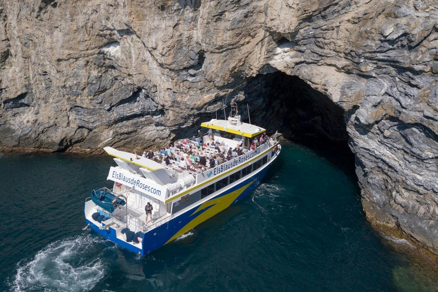 Un bateau transportant des passagers s'approche d'une grotte rocheuse sur le littoral ; on peut lire « ElsBlausdeRoses.com » sur son flanc.