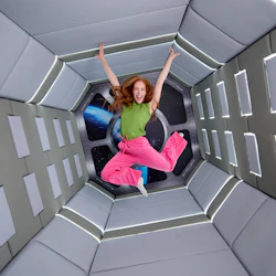 En person med långt hår, en grön topp och rosa byxor, hoppar in i ett futuristiskt, vadderat rum med ett rymdfönster.