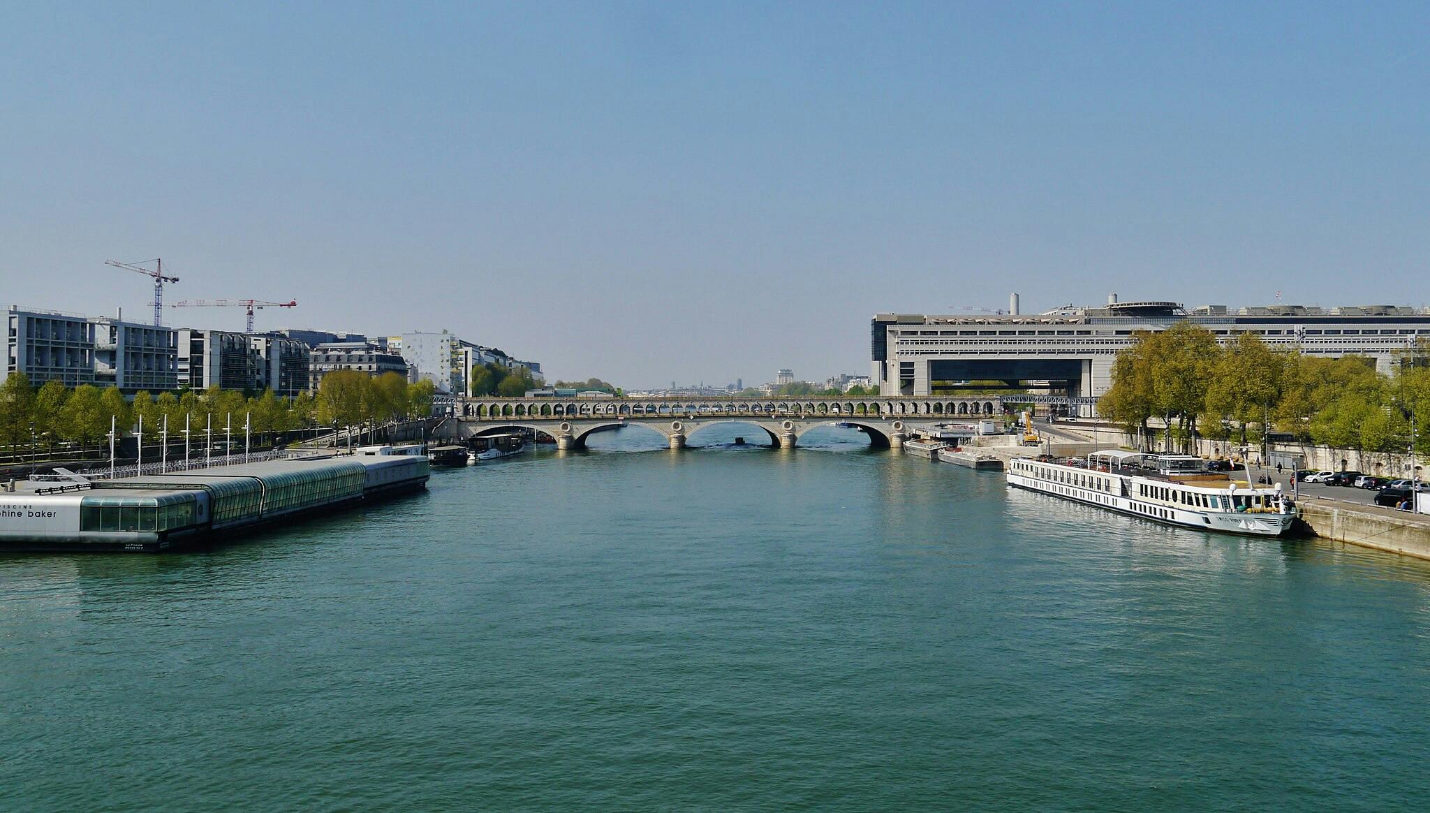 Ευρύ ποτάμι με βάρκες αγκυροβολημένες κατά μήκος των ακτών, μια πέτρινη γέφυρα που εκτείνεται στο νερό και σύγχρονα κτίρια και στις δύο πλευρές κάτω από έναν καθαρό ουρανό.