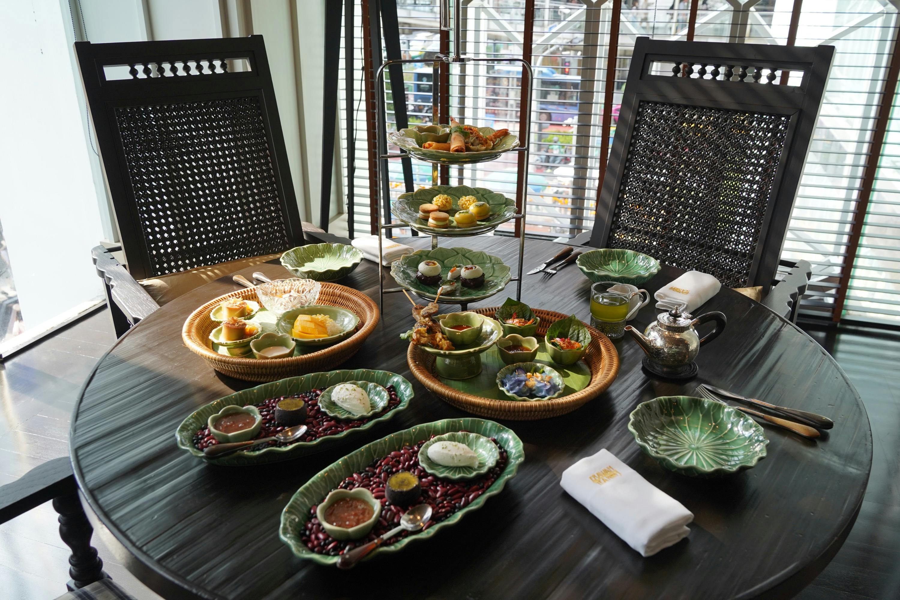 A dining table set with a variety of food items on green plates and a three-tiered serving stand, with chairs and a window behind.