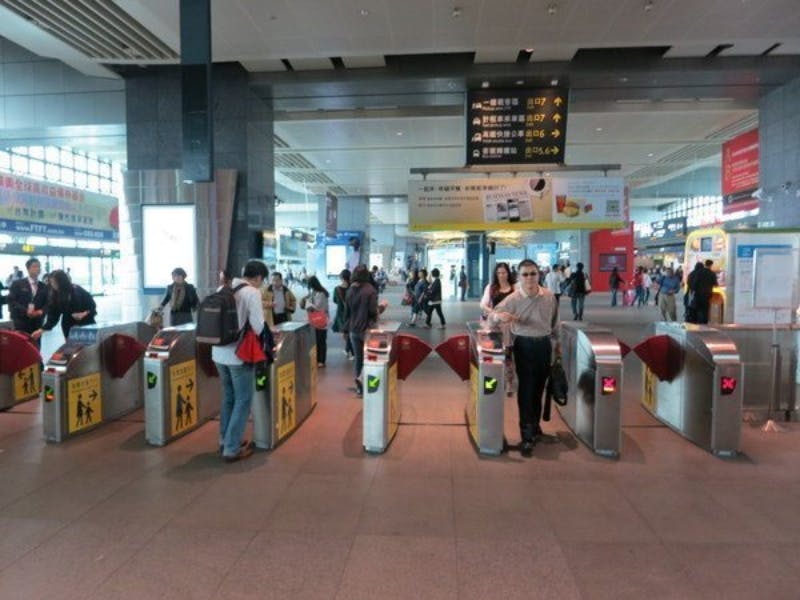 Άτομα που περνούν μέσα από περιστροφικές πύλες σε σιδηροδρομικό σταθμό με πινακίδες και πίνακες πληροφοριών στο παρασκήνιο.