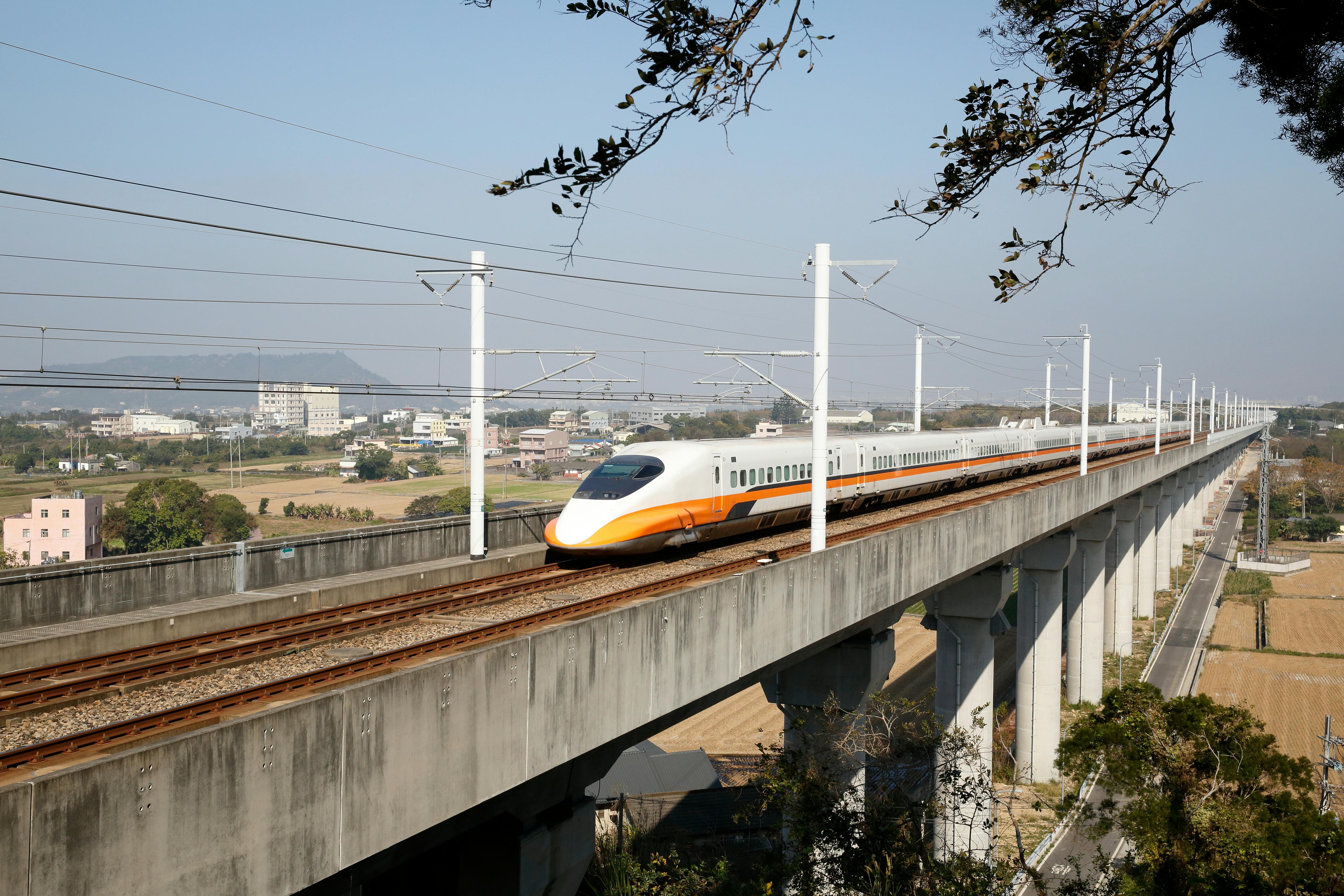 Ένα τρένο υψηλής ταχύτητας ταξιδεύει σε μια υπερυψωμένη διαδρομή πάνω από ένα αγροτικό τοπίο με διάσπαρτα κτίρια στο παρασκήνιο κάτω από έναν καθαρό ουρανό.