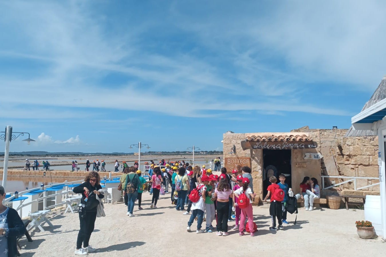 A group of people, including children with backpacks, gathered outdoors near a stone building under a partly cloudy sky.