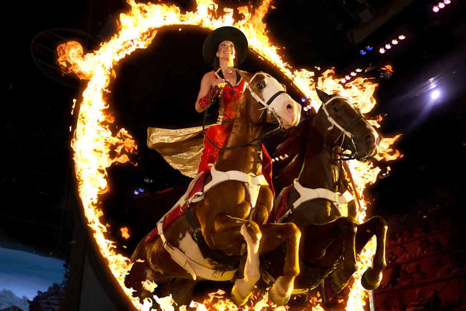 A performer on horseback jumping through a flaming ring, wearing a red outfit and a black hat, under a dark background.