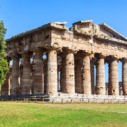 Forntida stentempel med höga pelare under en blå himmel, omgivet av ett gräsbevuxet område och ett stort träd till vänster.