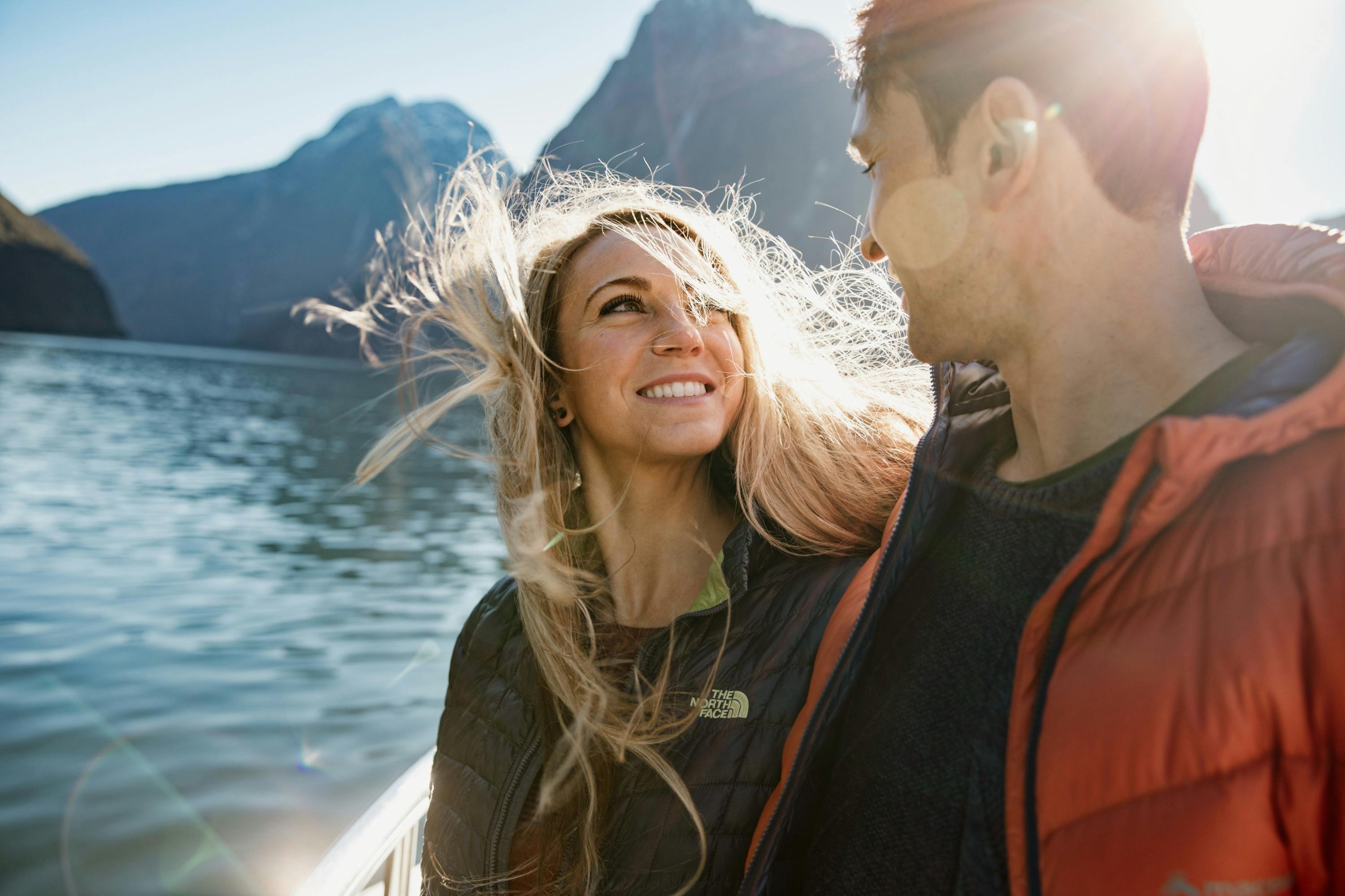 A woman and a man smiling at each other on a boat, surrounded by water and mountains on a sunny day.