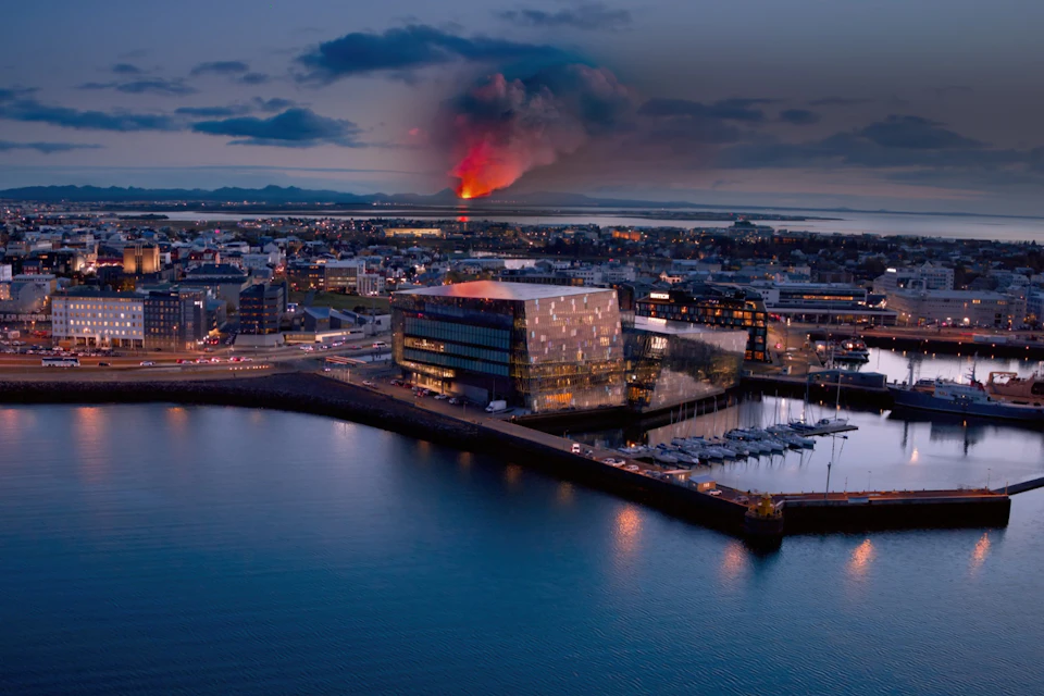 Aerial view of a city waterfront with a modern glass building, marina, and a distant volcano erupting under a cloudy sky.