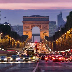 Utsikt över Triumfbågen i skymningen, med livlig trafik på Champs-Élysées kantad av träd och upplysta gatubelysning.