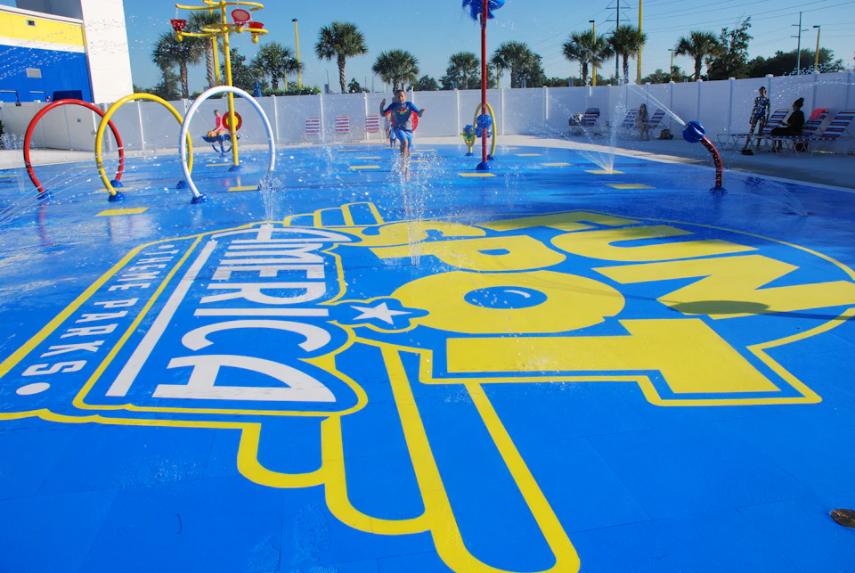 Child running through a colorful splash pad with fountains, featuring the words "Fun Spot America" on the blue surface.