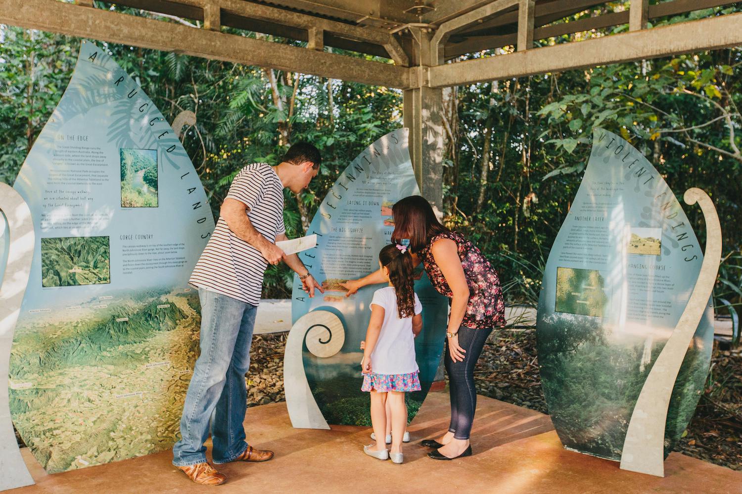 A man, woman, and child explore an educational outdoor exhibit in a forested area, reading informational panels.